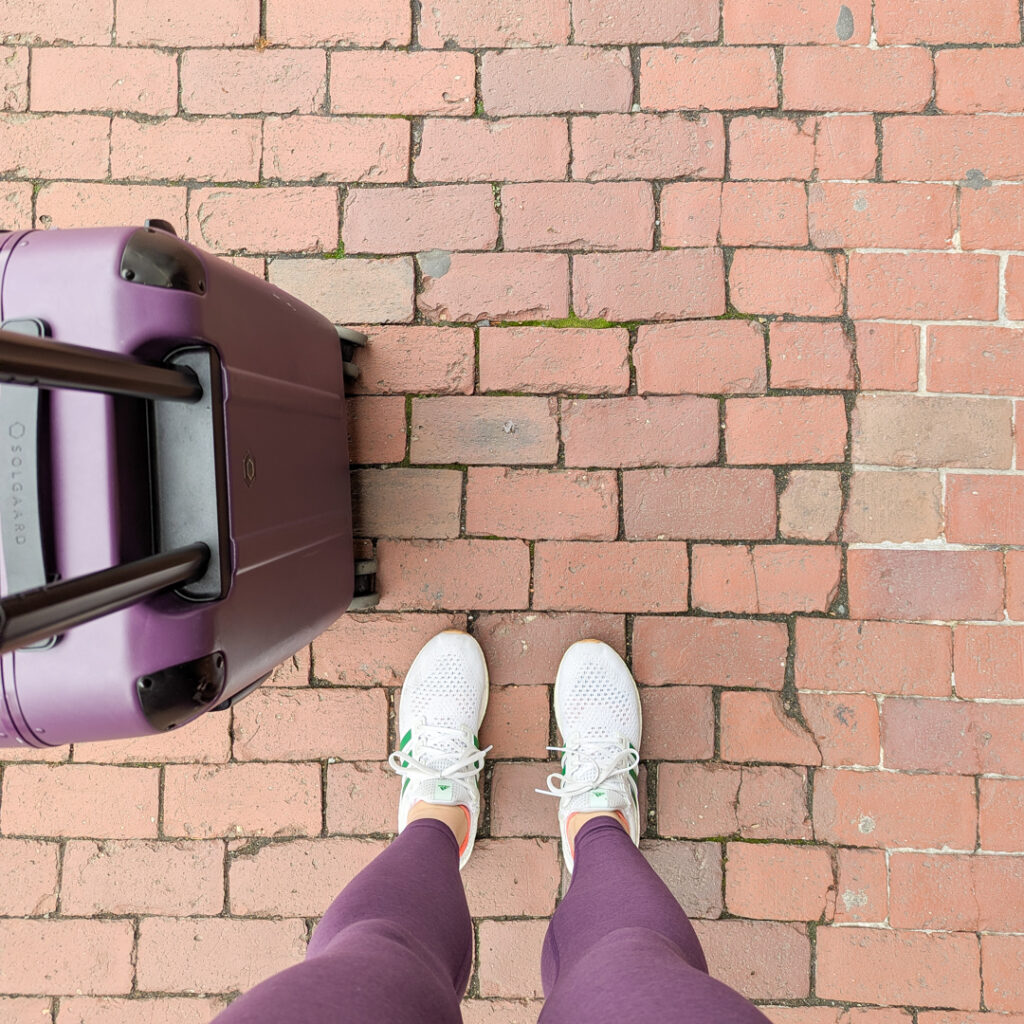 Looking down at a cobblestone sidewalk showing white shoes, purple leggings, and a purple rolling suitcase. 